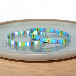 Könnte beinhalten: Zwei bunte Perlenarmbänder in Blau, Grün, Weiß und Gold. Die Armbänder liegen übereinander auf einem grauen Teller.
