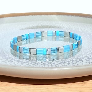 May include: A blue and silver beaded bracelet with a stretchy band. The bracelet is made of small, square beads in alternating colors.