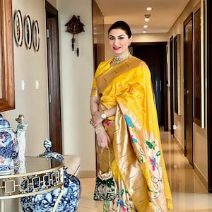 May include: A woman wearing a yellow and gold saree with a floral pattern. She is standing in a hallway with a decorative clock on the wall behind her.