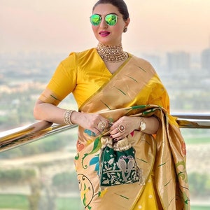 May include: A woman wearing a yellow saree with a green and gold embroidered clutch. She is wearing a gold necklace and sunglasses.