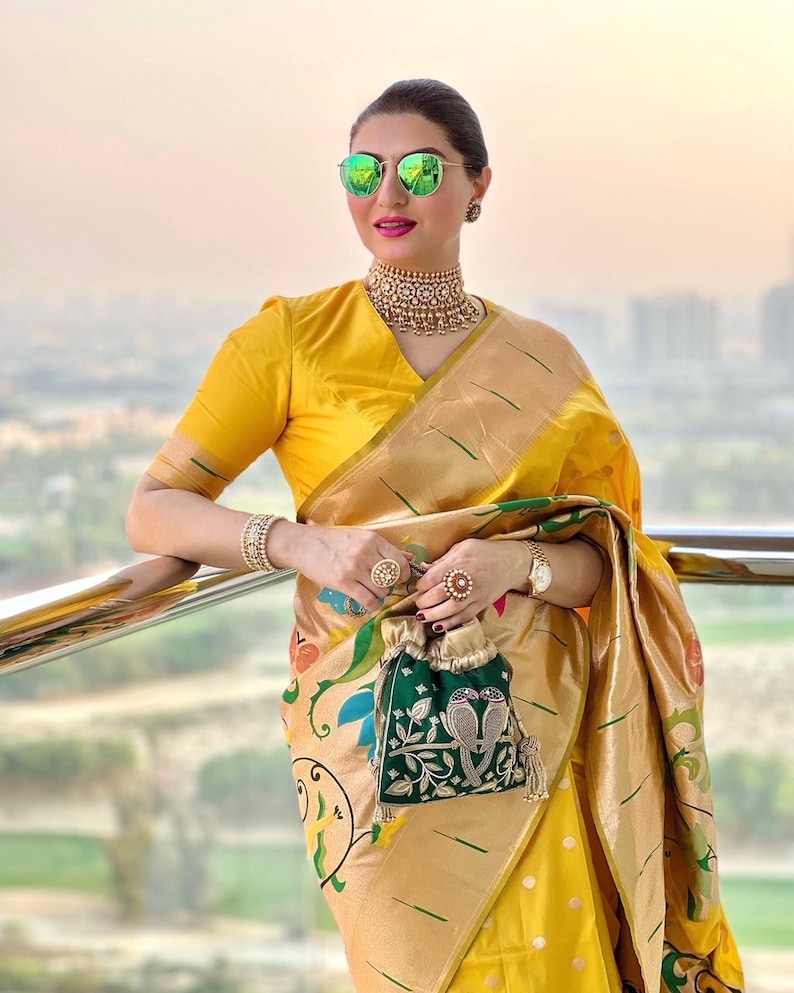 May include: A woman wearing a yellow saree with a green and gold embroidered clutch. She is wearing a gold necklace and sunglasses.