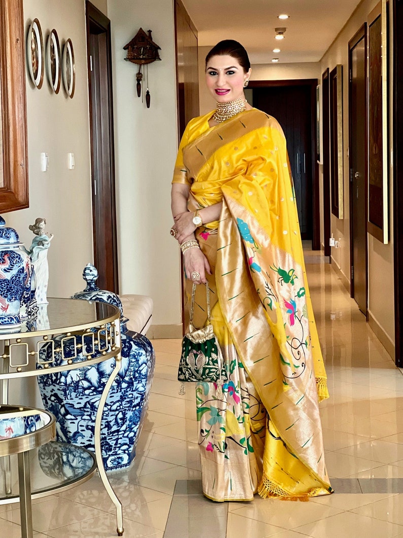 May include: A woman wearing a yellow and gold saree with a floral pattern. She is standing in a hallway with a decorative clock on the wall behind her.