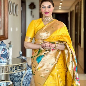 May include: A woman wearing a yellow and gold saree with a floral pattern. She is wearing a gold necklace and bracelets. The saree has a gold border and a green stripe.