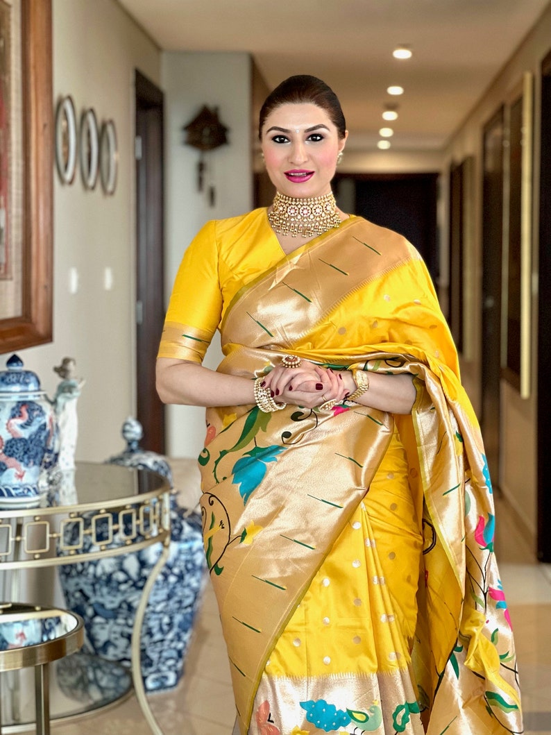 May include: A woman wearing a yellow and gold saree with a floral pattern. She is wearing a gold necklace and bracelets. The saree has a gold border and a green stripe.