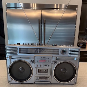 May include: A silver Rising brand portable boombox radio and cassette player featuring two large circular speakers with black mesh grilles, a central cassette deck, a radio frequency tuner display, multiple control knobs, and two long metal antennas.