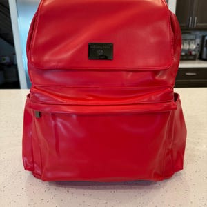 Könnte beinhalten: Ein leuchtend roter Rucksack mit glatter Oberfläche, ausgestattet mit einer großen oberen Klappe und einer Vordertasche. Eine rechteckige schwarze Metallplakette mit dem Text "Fawn Design" ist mittig auf der Klappe angebracht. Der Rucksack hat Seitentaschen.