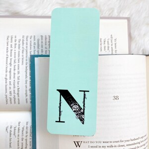 May include: A light blue bookmark with a black letter N decorated with vines and flowers. The bookmark is on top of a book with the page number 38 visible.