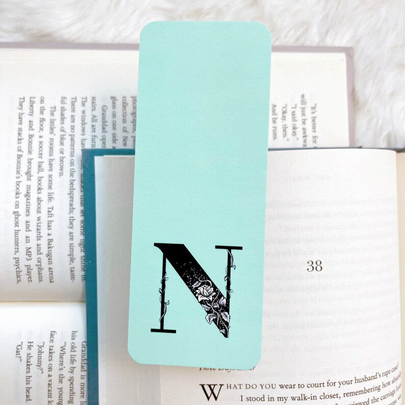 May include: A light blue bookmark with a black letter N decorated with vines and flowers. The bookmark is on top of a book with the page number 38 visible.