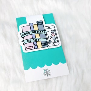 K&ouml;nnte beinhalten: Ein t&uuml;rkisfarbenes Lesezeichen mit einem wei&szlig;en Aufkleber, der einen Stapel bunter B&uuml;cher und ein Telefon mit dem Text "Booktok Made Me Do It" zeigt. Der Aufkleber enth&auml;lt auch eine Sprechblase mit dem Text "My Secret Copy".