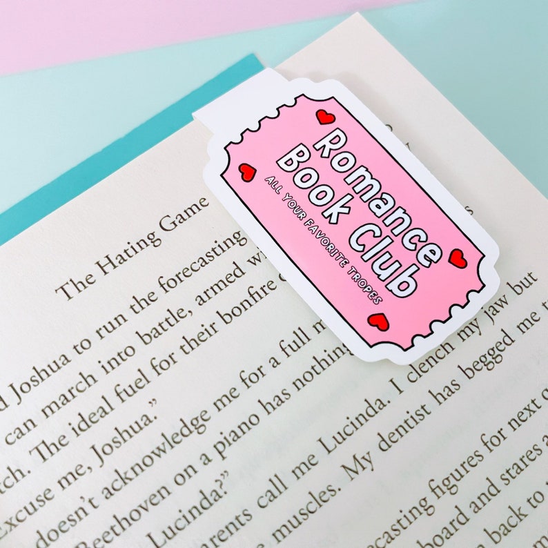 May include: Pink bookmark with a white border and red hearts. The bookmark has the text "Romance Book Club" and "All Your Favourite Tropes".