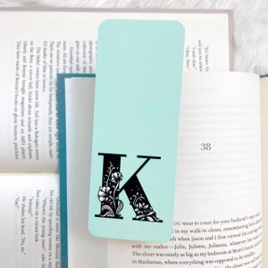 May include: A light blue bookmark with a black letter K decorated with white flowers. The bookmark has the number 38 printed on it.