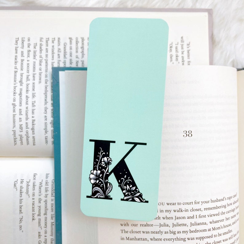 May include: A light blue bookmark with a black letter K decorated with white flowers. The bookmark has the number 38 printed on it.