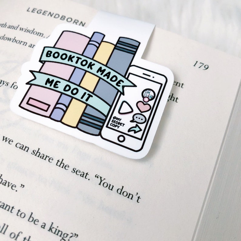 K&ouml;nnte beinhalten: Ein wei&szlig;es Lesezeichen mit einer bunten Illustration eines Stapels B&uuml;cher und eines Smartphones. Der Text auf dem Lesezeichen lautet "Booktok Made Me Do It".