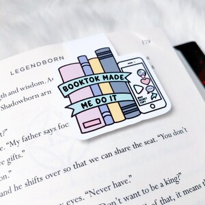 K&ouml;nnte beinhalten: Ein bunter Aufkleber mit einem Stapel B&uuml;cher und einem Telefon mit dem Text "Booktok Made Me Do It" auf einem blauen Banner.
