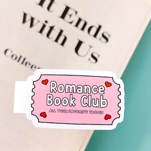 May include: Pink and white sticker in the shape of a ticket. It says "Romance Book Club" with hearts on either side. The text below says "All Your Favourite Tropes".
