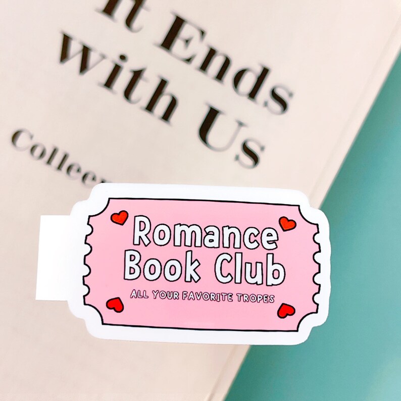 May include: Pink and white sticker in the shape of a ticket. It says "Romance Book Club" with hearts on either side. The text below says "All Your Favourite Tropes".