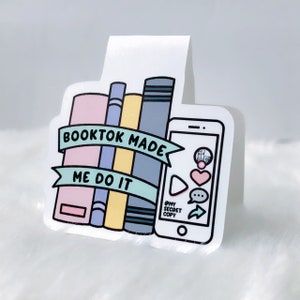 K&ouml;nnte beinhalten: Ein wei&szlig;es Lesezeichen mit einer bunten Illustration eines B&uuml;cherstapels und eines Telefons. Der Text auf dem Lesezeichen lautet "Booktok Made Me Do It" und "@mysecretcopy".