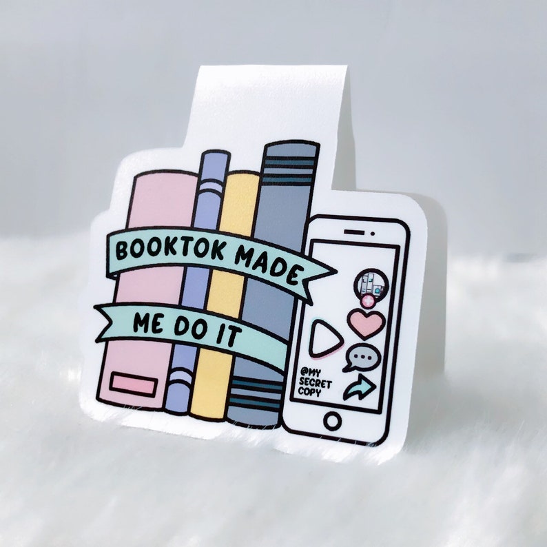 K&ouml;nnte beinhalten: Ein wei&szlig;es Lesezeichen mit einer bunten Illustration eines B&uuml;cherstapels und eines Telefons. Der Text auf dem Lesezeichen lautet "Booktok Made Me Do It" und "@mysecretcopy".