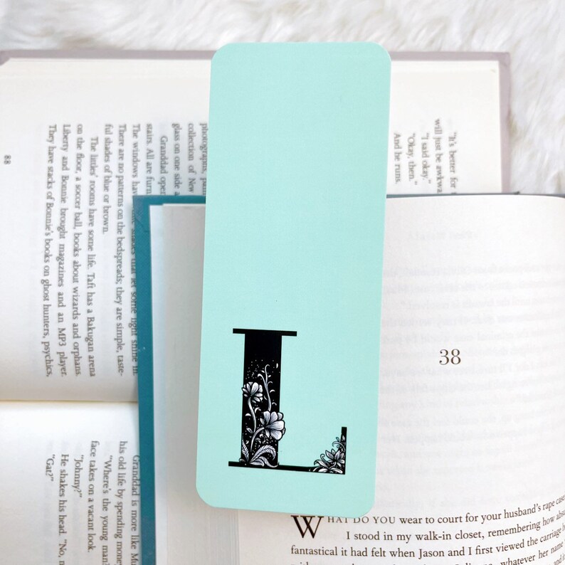 May include: A light blue bookmark with a black letter L decorated with floral designs. The bookmark is placed on top of an open book with the page number 38 visible.