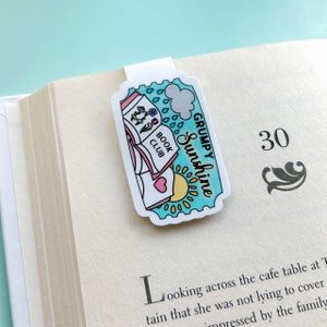 Può includere: Un segnalibro blu e bianco con un libro, fiori, un sole e una nuvola con gocce di pioggia. Il testo "GRUMPY Sunshine" e "BOOK CLUB" sono sul segnalibro.