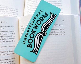May include: A turquoise blue bookmark with a black and white illustration of a book. The bookmark has the text "Unprofessional Bookworm" in black letters.