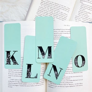 May include: Set of five turquoise blue bookmarks with black floral illustrations and the letters K, L, M, N, and O. The bookmarks are on a book with white pages.