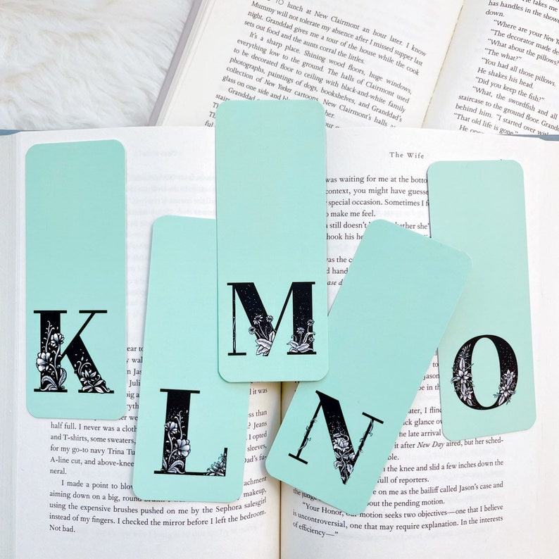 May include: Set of five turquoise blue bookmarks with black floral illustrations and the letters K, L, M, N, and O. The bookmarks are on a book with white pages.
