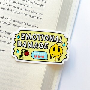 May include: Yellow sticker with the text "Emotional Damage" in black letters. A yellow smiley face with a dripping yellow substance is on the right side of the sticker. A blue speech bubble with the text "book club" and two broken red hearts is on the left side of the sticker.