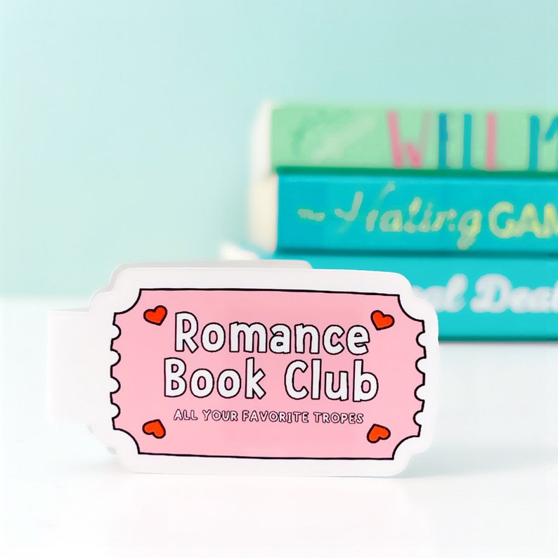 May include: A pink ticket-shaped bookmark with the text "Romance Book Club" and the words "All Your Favorite Tropes" in smaller font. There are small red hearts around the text.