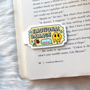 May include: A yellow and white bookmark with the text "Emotional Damage" and a cartoon smiley face. The bookmark is shaped like a ticket and has a small speech bubble with the text "Book Club".
