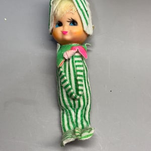 May include: A small doll with blonde hair, blue eyes, and pink lips, wearing a green and white striped fabric outfit that covers the body and head. The doll features a green collar with a small pink accent piece at the neck area.