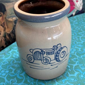 May include: A cylindrical stoneware crock with a beige body and a dark brown interior. The rim is accented with a blue band, and the front features a blue line-art illustration of a tractor. The vessel has a smooth, glazed finish.