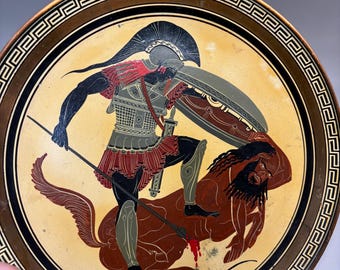 May include: A round decorative plate featuring a black-figure style illustration of a warrior in a helmet and armor holding a spear and shield while standing over a fallen centaur. The border displays a classic Greek key pattern in black and gold.