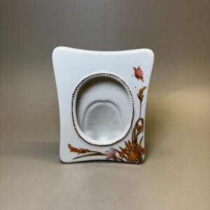 Puede incluir: Un marco de fotos de cerámica blanca con forma rectangular ensanchada y una abertura central ovalada. El marco presenta un borde dorado alrededor de la abertura y un diseño pintado de flores de iris naranjas y una pequeña mariposa rosa.