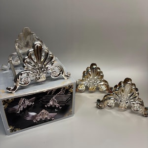May include: A set of three silver-toned metal cutlery rests featuring a decorative shell-like motif with ornate scrollwork. One rest is positioned on top of its original gray and black cardboard box, while two additional rests sit to the right.