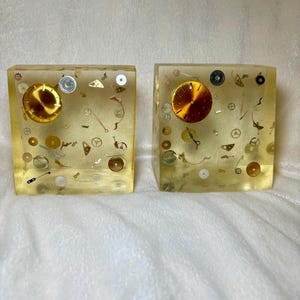 May include: Two square blocks made of clear, pale yellow resin, each containing embedded metallic watch parts and gears. A circular, gold-colored watch face dial is positioned in the upper left corner of each block, surrounded by various small silver and brass clock components.