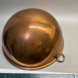 May include: A polished copper mixing bowl with a smooth, rounded dome shape. The bowl features a single brass ring handle attached to the side rim, allowing for easy hanging or handling. The surface is highly reflective with a warm metallic finish.