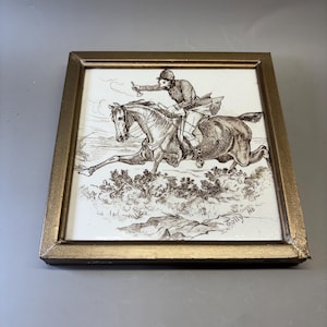 May include: A square ceramic tile framed in gold, featuring a brown ink illustration of a person riding a horse while blowing a hunting horn. The scene depicts a fox hunt in a field, with the text Tally Ho written in the bottom right corner.
