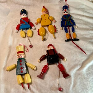 May include: Five wooden jumping jack ornaments with pull strings. The figures include a soldier in red and blue, a yellow clown, a blue soldier with a mustache, a green figure in a tall hat, and a figure in black overalls with red boots.
