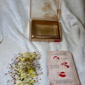 May include: A clear plastic rectangular box with a gold-toned textured frame, a collection of small glass ampoules filled with yellow liquid, and an instructional paper titled HOW TO USE NIPS with black text and illustrations showing usage steps.