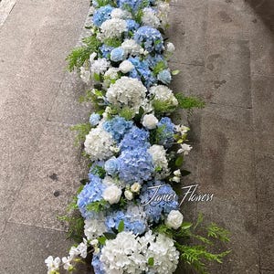 Könnte beinhalten: Ein langes Blumenarrangement mit weißen und blauen Hortensien, kleinen weißen Rosen und grünem Laub. Das Arrangement wird auf einer grauen Oberfläche präsentiert und enthält den Text "James Flowers".