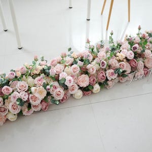 May include: A long floral arrangement featuring pink and cream roses and green foliage. The arrangement is displayed on a white surface. The text "James Flowers" is visible in the lower right corner.