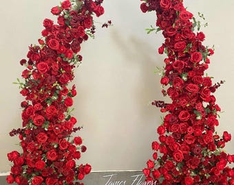 May include: Two floral archways composed of vibrant red roses and greenery. The arches are densely packed with blooms, creating a full and lush appearance. The text "James Flowers" is visible at the bottom.