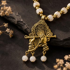 May include: A gold-toned metal pendant necklace featuring a detailed depiction of hands playing a flute, accented with a circular filigree design. The necklace chain consists of smooth white beads separated by small gold-toned metal spacers.