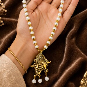 May include: A necklace featuring a string of white spherical beads separated by small gold-toned spacers. The pendant is a gold-toned metal design depicting two hands holding a flute, with three small white beads dangling from the bottom of the piece.