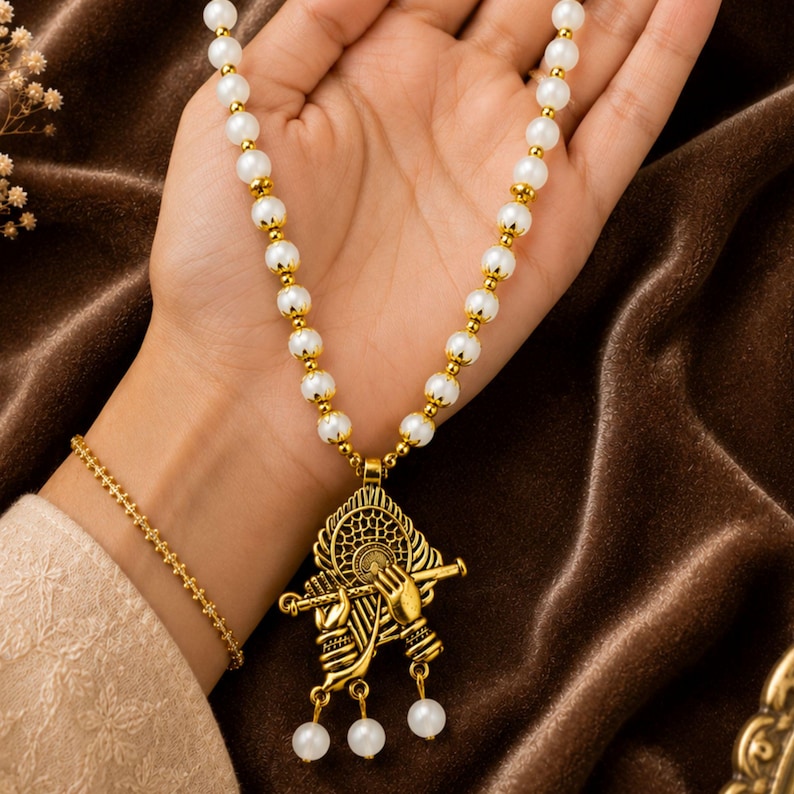 May include: A necklace featuring a string of white spherical beads separated by small gold-toned spacers. The pendant is a gold-toned metal design depicting two hands holding a flute, with three small white beads dangling from the bottom of the piece.