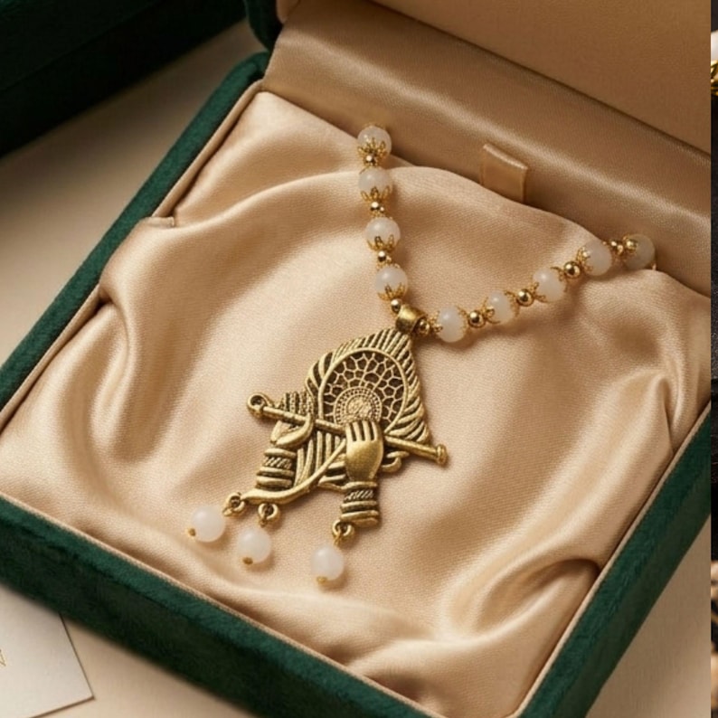 May include: A necklace featuring a gold-toned metal pendant shaped like a peacock feather with a flute and hand motif. The chain consists of white spherical beads interspersed with small gold-toned metal spacers, ending in three dangling white beads.