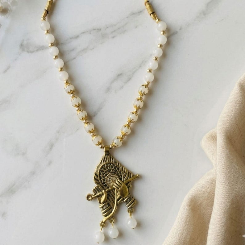 May include: A necklace featuring a string of white beads separated by small gold-toned metal spacers. The pendant is a gold-toned metal charm depicting a figure playing a flute, accented with three small white beads hanging from the bottom.