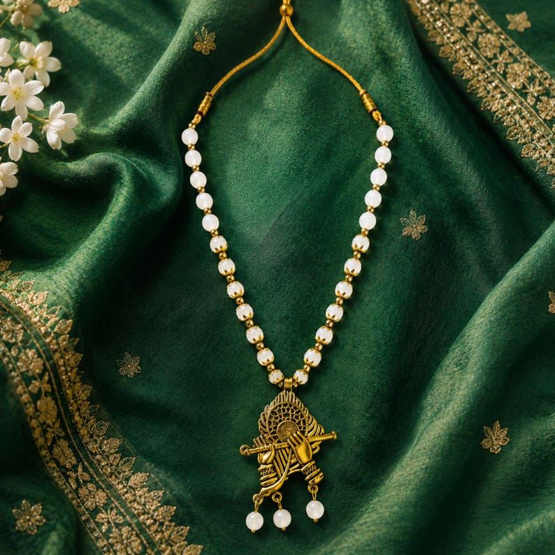 May include: A necklace featuring a series of round, translucent white beads strung on a gold-toned cord. The pendant is a gold-toned metal charm depicting a pair of hands holding a flute, with three small white beads dangling from the bottom.