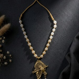 May include: A necklace featuring a series of smooth, round, off-white beads interspersed with small, ornate gold-toned metal spacers. The pendant is a detailed gold-toned metal design depicting a hand holding a flute against a circular frame.